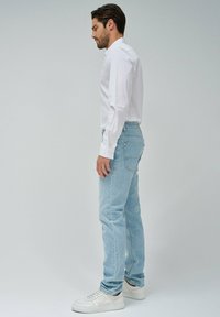 Jeans de un azul claro con un corte clásico, material de mezclilla, con un ligero desgaste y detalles en el bolsillo trasero, combinados con una camisa blanca de botones y zapatillas blancas.