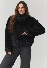 Czarna kurtka z sztucznego futra, krótka, z wysokim kołnierzem, długimi rękawami i miękką teksturą. Noszona z ciemnymi dżinsami.