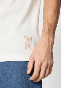 T-shirt en coton blanc avec une étiquette crème portant un logo et le texte "Italia" en bas à gauche ; porté avec un jean en denim bleu.