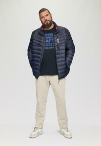 Donkerblauwe pufferjack met gestikt patroon, zwart graphic t-shirt, beige broek en lichte sneakers. Eenvoudig design, praktische kenmerken.