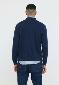 Sweatshirt bleu marine avec un col rond, des poignets et un ourlet côtelés, fabriqué en mélange de coton doux. Porté par-dessus une chemise à col bleu clair.