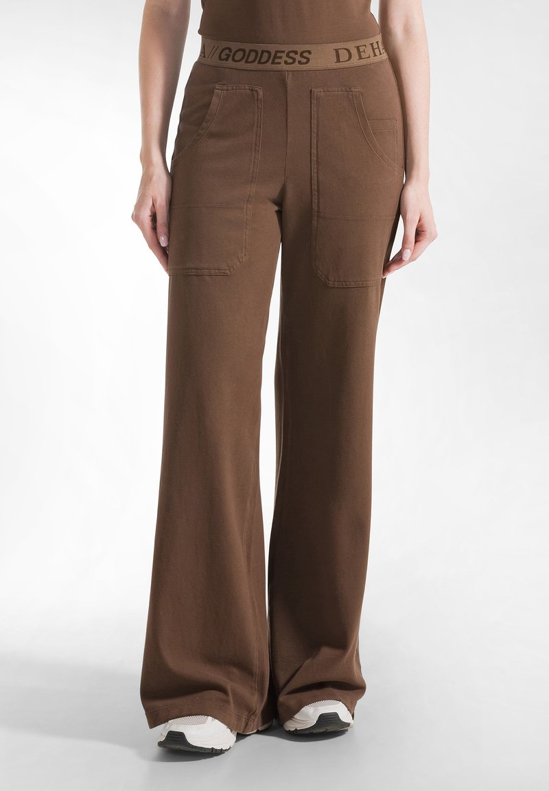 Bruine wijde broek met een aansluitende tailleband met reliëftekst, twee voorkantzakken en een zachte katoenstructuur. In combinatie met witte sneakers.