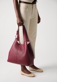 Stor, burgunderlæder tote bag med en tekstureret overflade og blød, slasket silhuet. Har kontrasterende cremede accenter og en detalje med guldfinish.