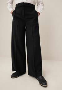 Schwarze, weit geschnittene Hosen mit glatter Textur, hoher Taille und vorderen Falten, kombiniert mit schwarzen, fransigen Slippers.