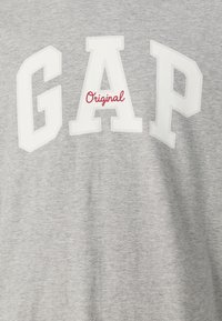 T-shirt en coton gris avec de grandes lettres blanches en bloc épelant "GAP" et le mot "Original" en cursive rouge, texture douce et design décontracté.