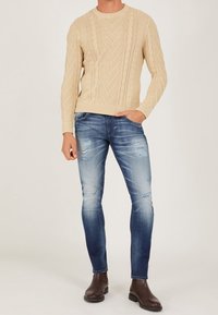 Man som bär en beige kabelstickad tröja, slim fit slitna blå jeans och mörkbruna läderankelstövlar, står med en hand i fickan.