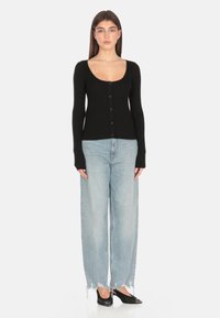 Cardigan nero a costine con scollatura a barca e chiusura a bottone, abbinato a jeans baggy azzurri chiari con orli sfrangiati e scarpe nere.