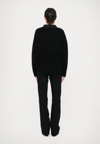 Schwarzer, oversized Strickpullover mit Rundhalsausschnitt, strukturiertem Stoff und überschnittenen Schultern; kombiniert mit schwarzen, eng anliegenden Hosen, von hinten gezeigt.