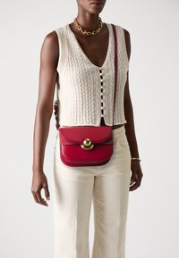Borsa a tracolla in pelle rossa con un clasp circolare dorato e cuciture a contrasto. Indossata con un top lavorato a maglia color crema e pantaloni chiari.