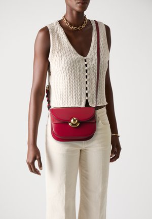 Red leather crossbody bag with a gold circular clasp and contrasting stitching. Worn with a cream knitted top and light-colored trousers.