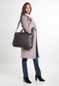 Borsa a mano in pelle marrone con due manici e tracolla removibile. Superficie texture, forma rettangolare, con tasca frontale e branding discreto.