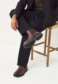 Hnedé kožené členkové topánky s elastickými bočnými panelmi, plochými čiernymi gumovými podrážkami a štruktúrovaným povrchom, nosené s čiernymi nohavicami, sediace na drevenej stoličke.