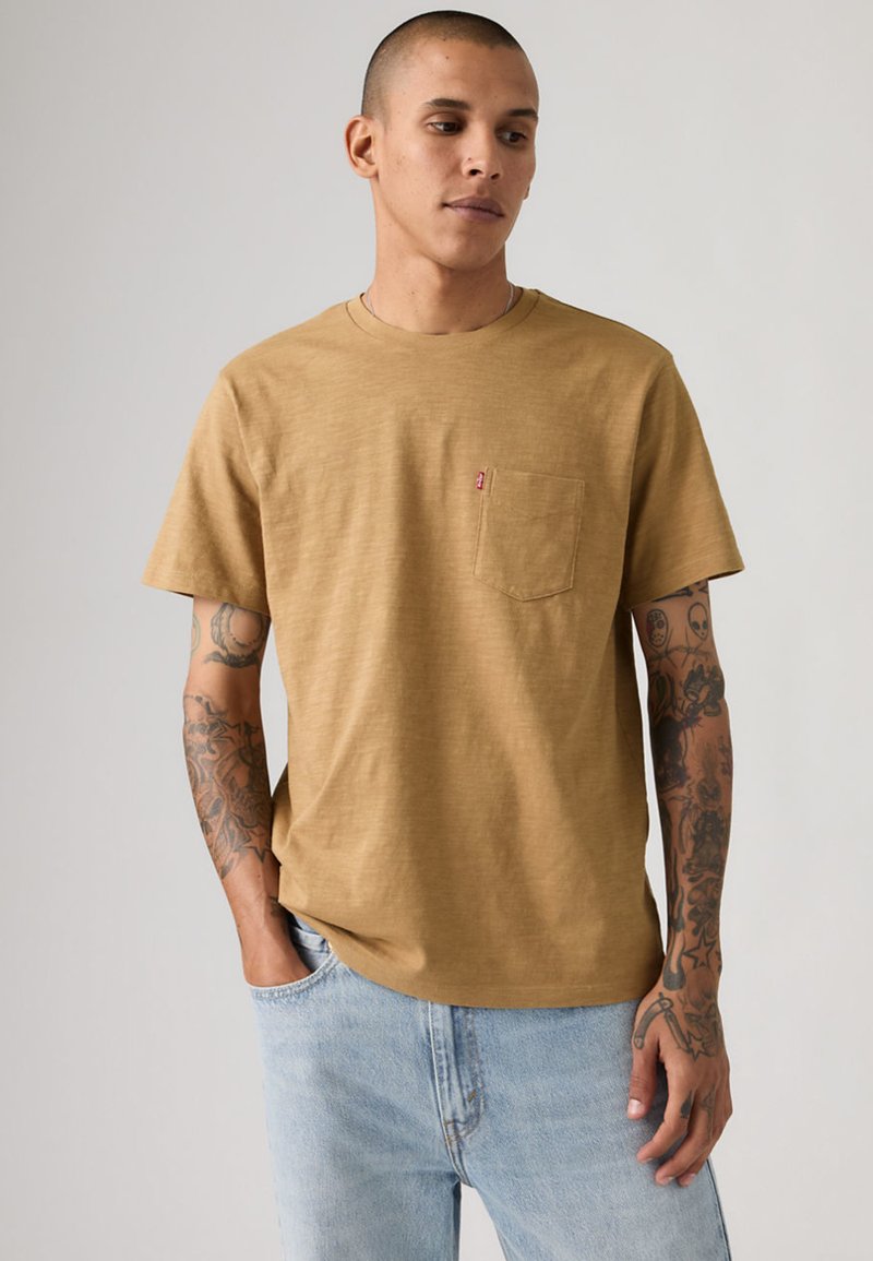 Man with tattoos wearing mustard yellow t-shirt with chest pocket and light blue jeans, standing with one hand in pocket, looking down.