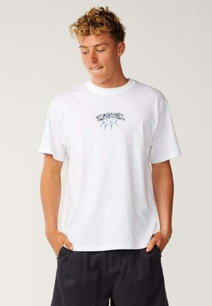 Jeune homme aux cheveux bouclés portant un t-shirt blanc Rip Curl et un short noir, debout les mains dans les poches, regardant vers le bas.