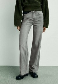 Sive široke traperice z neobdelanim robom, v kombinaciji z zelenim teksturiranim puloverjem in črnimi čevlji. Osredotoča se na sproščen stil in nevtralne barve.