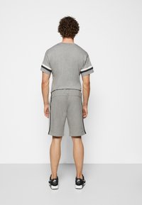 Muž s kudrnatými vlasy, oblečený v šedé košili s krátkým rukávem a odpovídajících šortkách se postranními pruhy, v černobílých botách Nike Air, stojící zády.