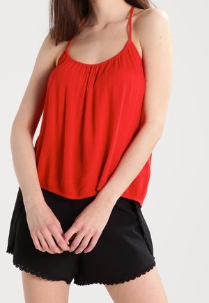 Rotes ärmelloses Oberteil mit gerafftem Ausschnitt und dünnen Trägern, kombiniert mit schwarzen Shorts mit gewelltem Spitzenbesatz. Glatte Stoffstruktur.