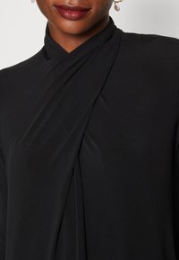 Musta kääntöyöpaita, joka on valmistettu sileästä kankaasta, jossa on risteävä muotoilu kaula-aukossa ja elegantti laskostus sivuilla.