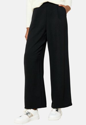 Pantalons larges noirs en tissu doux, dotés d'une taille haute et de poches latérales, associés à des baskets blanches avec des accents métalliques.