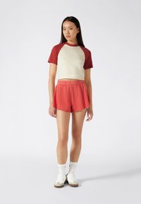 Short-sleeve, beige and red color-blocked t-shirt paired with red athletic shorts, featuring an elastic waistband and curved hem. White sneakers with gray accents.