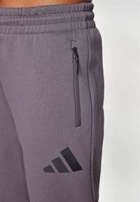 Grå idrettsjoggere med elastisk midjebånd, glidelåslomme og svart Adidas-logo. Glatt tekstur og tettsittende design.