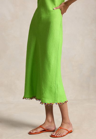 Nő, aki élénk zöld midi szoknyát visel többszínű gyöngyözött szegéllyel és narancssárga papucsot, kezét a csípőjén tartva áll.