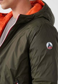 Giacca verde oliva con fodera arancione nel cappuccio, chiusura con zip e accento rosso. Presenta una patch con logo sulla spalla sinistra. Tessuto dalla texture liscia.