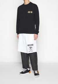 Černá mikina s metalickým logem "MOSCHINO", bílé šortky s potiskem "MOSCHINO MILANO", pruhované kalhoty a černé tenisky s oranžovými akcenty.