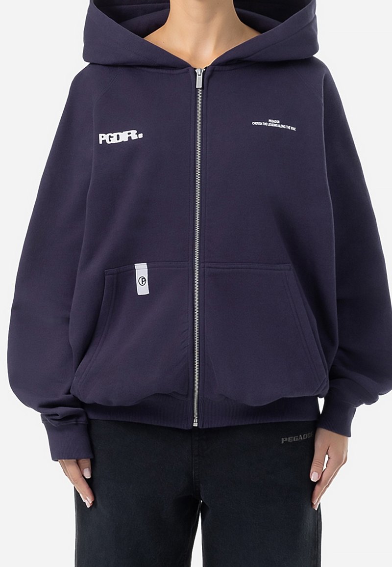 Personne portant un sweat à capuche bleu marine zippé avec des manches surdimensionnées et des poches avant, associé à un pantalon noir, sur un fond uni.