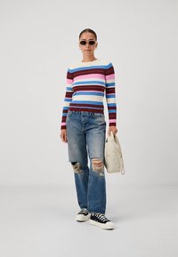 Pulover cu dungi multicolore în albastru, roz, roșu și crem, cu textură nervurată; asortat cu blugi drepți cu rupturi și adidași negri.