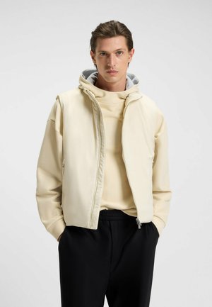 Light beige sleeveless jacket with zip closure, featuring a hood, layered over a soft cream sweater, paired with black pants. Smooth texture.