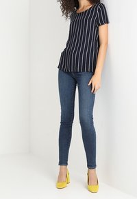 Frau trägt ein schwarzes Kurzarmoberteil mit weißen vertikalen Streifen, eng anliegende blaue Jeans und gelbe Blockabsatzschuhe, steht vor weißen Wänden.