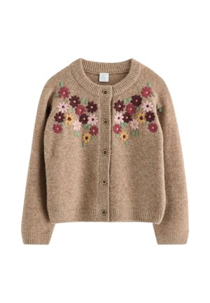 Smilende pige med krøllet hår iført en brun cardigan med blomsterprint, løse blå jeans og sorte sko poserer med armene udstrakt.