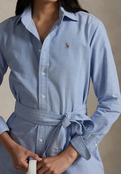 Moterys vilkinti šviesiai mėlyną marškinių tipo suknelę su diržu ir sagomis, laiko baltos rankinės dirželį, matomas polo žaidėjo logotipas ant krūtinės.