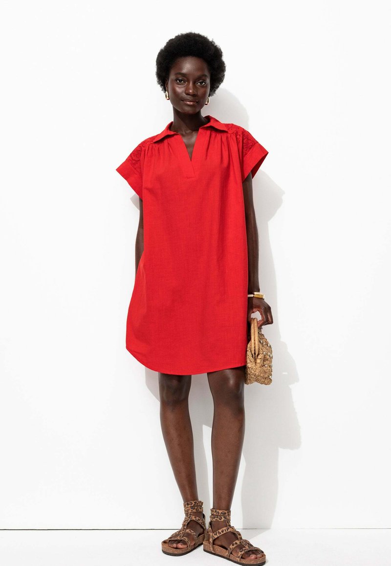 Robe en lin rouge avec col, manches courtes et décolleté en V. Accents texturés sur les manches. Associée à des sandales marron à imprimé léopard.