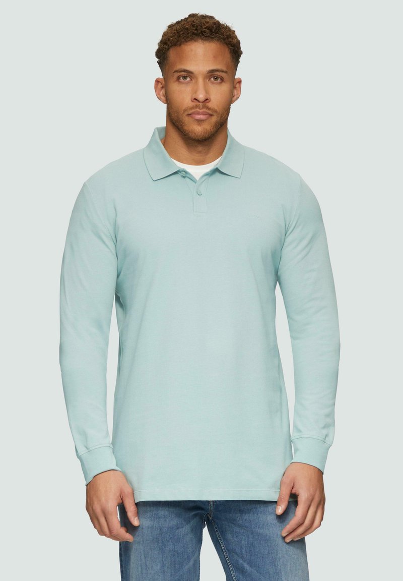 Polo-shirt à manches longues bleu clair avec un col, fabriqué en tissu doux. Présente un design minimaliste et un ourlet droit ; porté avec un jean bleu.