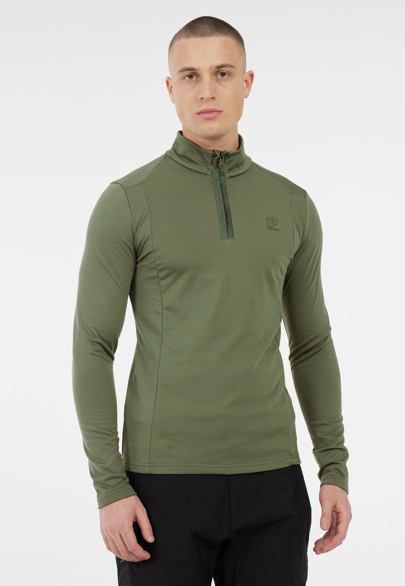 Giovane con capelli corti che indossa una maglietta a maniche lunghe verde oliva con zip e pantaloni neri, in piedi su uno sfondo bianco.