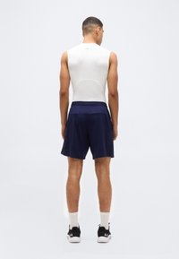 Άνδρας που στέκεται με την πλάτη, φοράει λευκό αμάνικο συμπιεστικό μπλουζάκι, ναυτικό αθλητικό σορτς, λευκές κάλτσες και μαύρα αθλητικά παπούτσια.