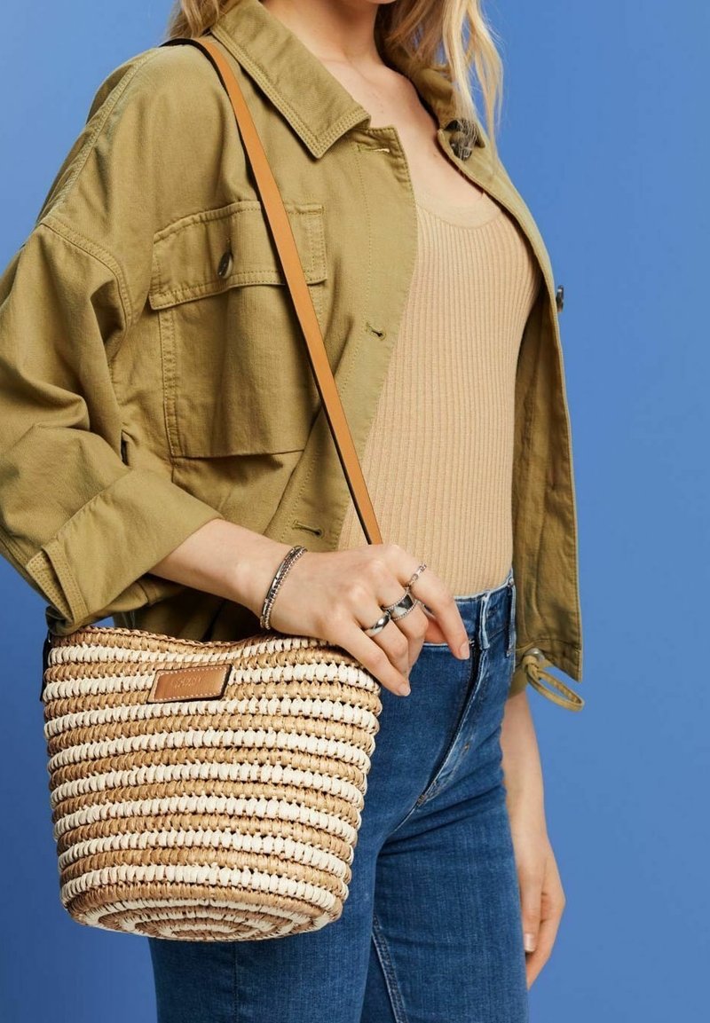 Femme portant une veste marron clair et un haut beige avec un jean bleu, tenant un sac bandoulière en paille tressée avec une lanière beige sur fond bleu.