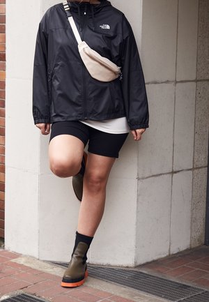 Persoon die een zwarte North Face jas, zwarte korte broek, beige crossbody tas en groene laarzen met oranje zolen draagt, staand op één been tegen een muur.