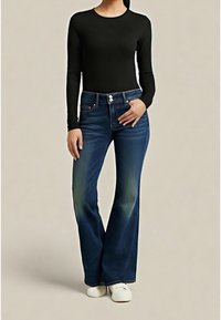 Personne portant un jean évasé bleu foncé avec fermeture à double bouton, un haut noir à manches longues et des chaussures blanches sans lacets, debout devant un fond beige.
