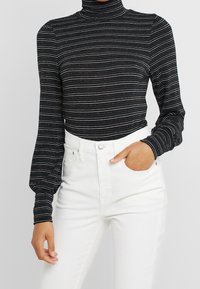 Haut à manches longues noir et métallique à rayures avec un col roulé haut, associé à un pantalon blanc taille haute. La texture semble côtelée.