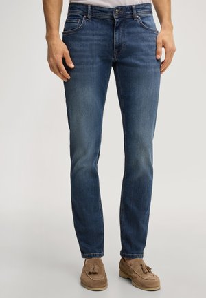 Mężczyzna w dopasowanych niebieskich dżinsach i brązowych zamszowych mokasynach z chwostami, stoi z rękami swobodnie opuszczonymi wzdłuż ciała.