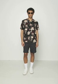 Kortermet svart skjorte med blomsterdesign, kombinert med mørke shorts og hvite joggesko. Modellen har på seg solbriller og hvite ankelsokker.