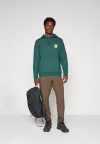 Man in groene hoodie en bruine broek, met een zwarte rugzak, staand tegen een effen lichtgrijze achtergrond.