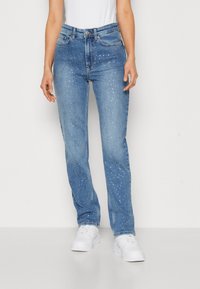 Jeans de mezclilla azul claro con un ajuste de pierna recta, con tachuelas plateadas esparcidas por la tela. Combinados con zapatillas blancas.