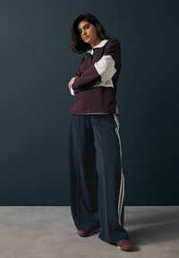 Polo à manches longues bordeaux et blanc associé à un pantalon large bleu marine avec des rayures blanches. Le mannequin se tient avec confiance devant un fond sombre.