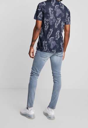 Chemise bleu marine à manches courtes avec un motif cachemire, associée à un jean bleu clair ajusté et des baskets blanches à semelle translucide.