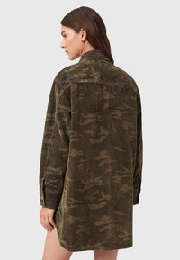 Frau mit braunen Haaren, die eine lange, braun-schwarze Cargohosenjacke mit Tarnmuster trägt und seitlich vor einem schlichten hellen Hintergrund steht.