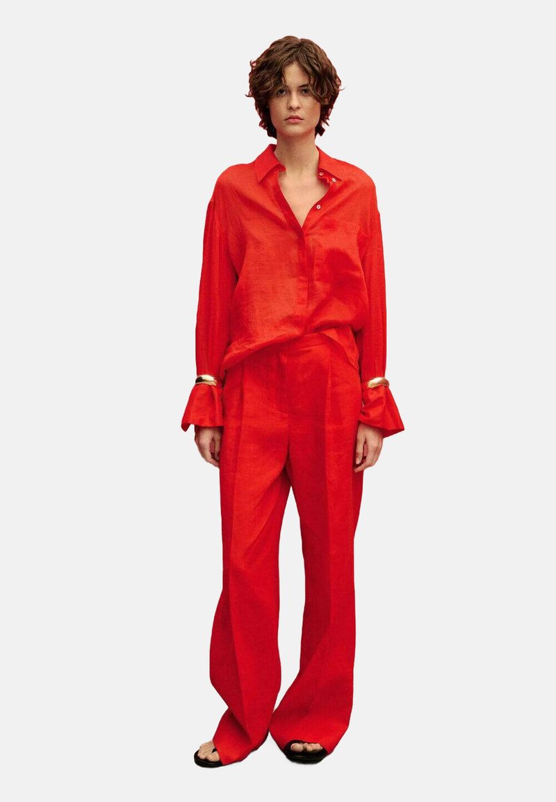 Chemise en lin rouge et pantalon ample, avec des manches longues et des poignets évasés, offrant une silhouette décontractée. Porté avec des sandales noires simples.